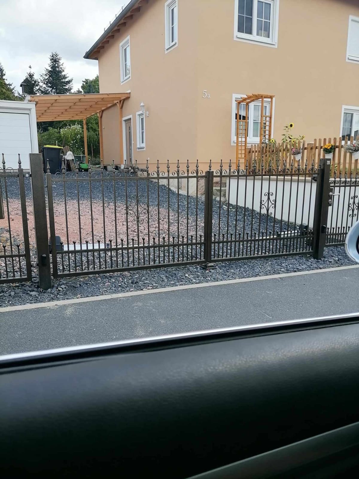 Czarna, metalowa brama wjazdowa z dekoracyjnymi elementami, widok zza szyby samochodu, dom z beżową elewacją w tle.