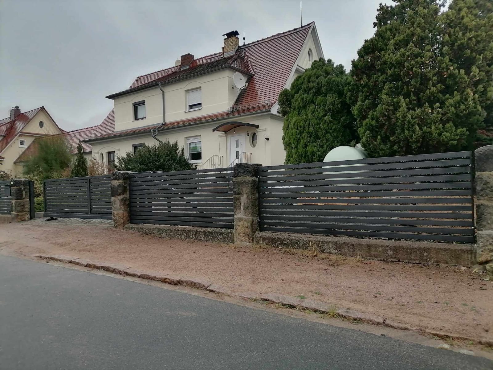 Nowoczesne ogrodzenie metalowe z poziomymi panelami w kolorze grafitowym, osadzone na kamiennych słupkach, na tle domu z czerwonym dachem i zielenią.