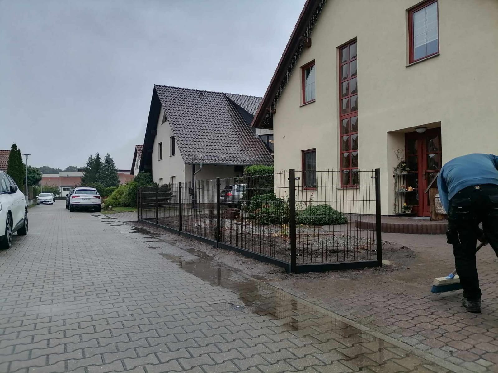 Nowo zamontowane, czarne ogrodzenie z paneli drucianych oddzielające posesję z domem jednorodzinnym od chodnika; pracownik zamiata bruk po instalacji.