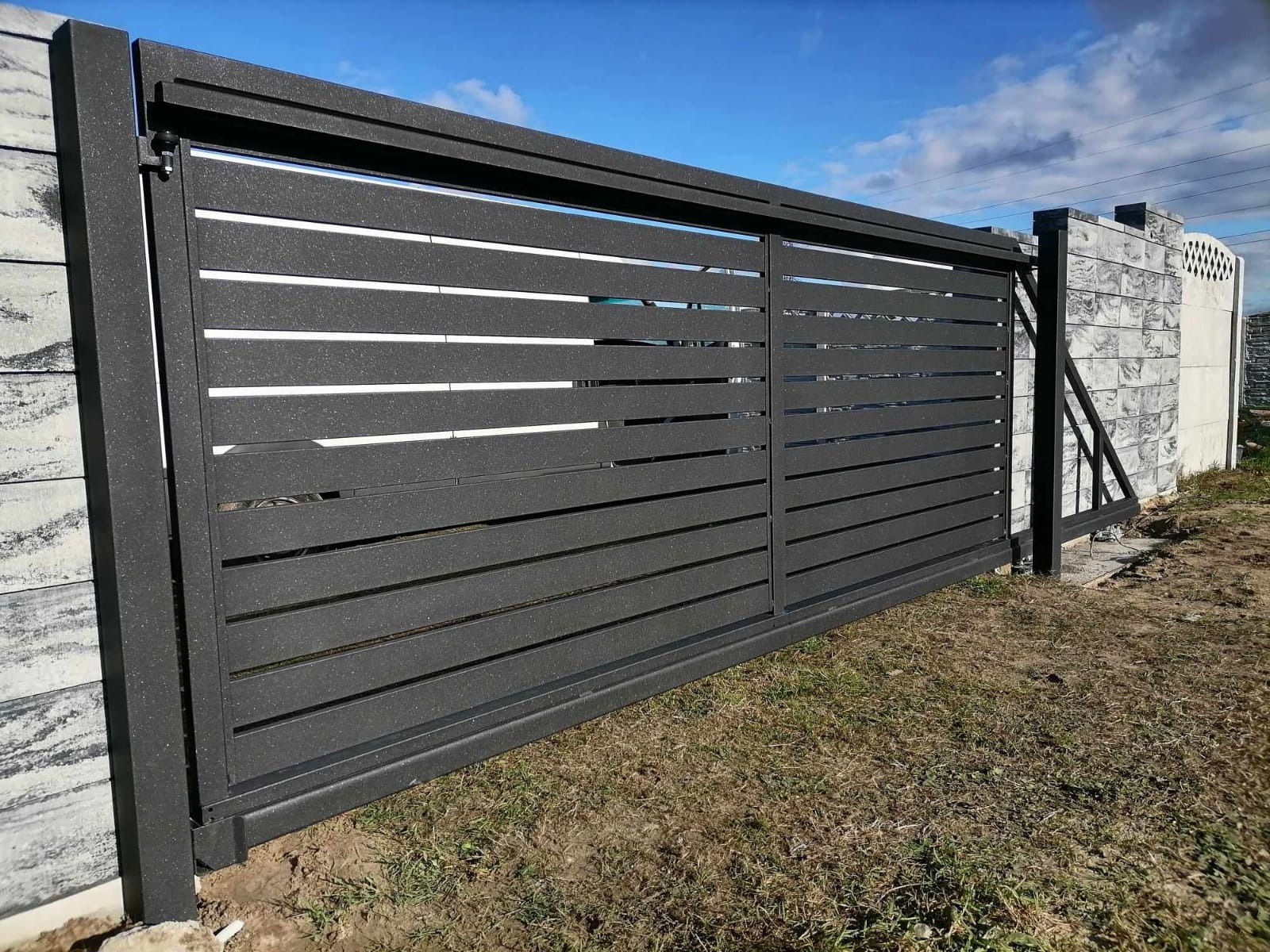 Przesuwna brama z poziomych, metalowych profili, antracytowa, zamontowana na posesji z betonowymi słupkami, widoczna trawa i błękitne niebo.