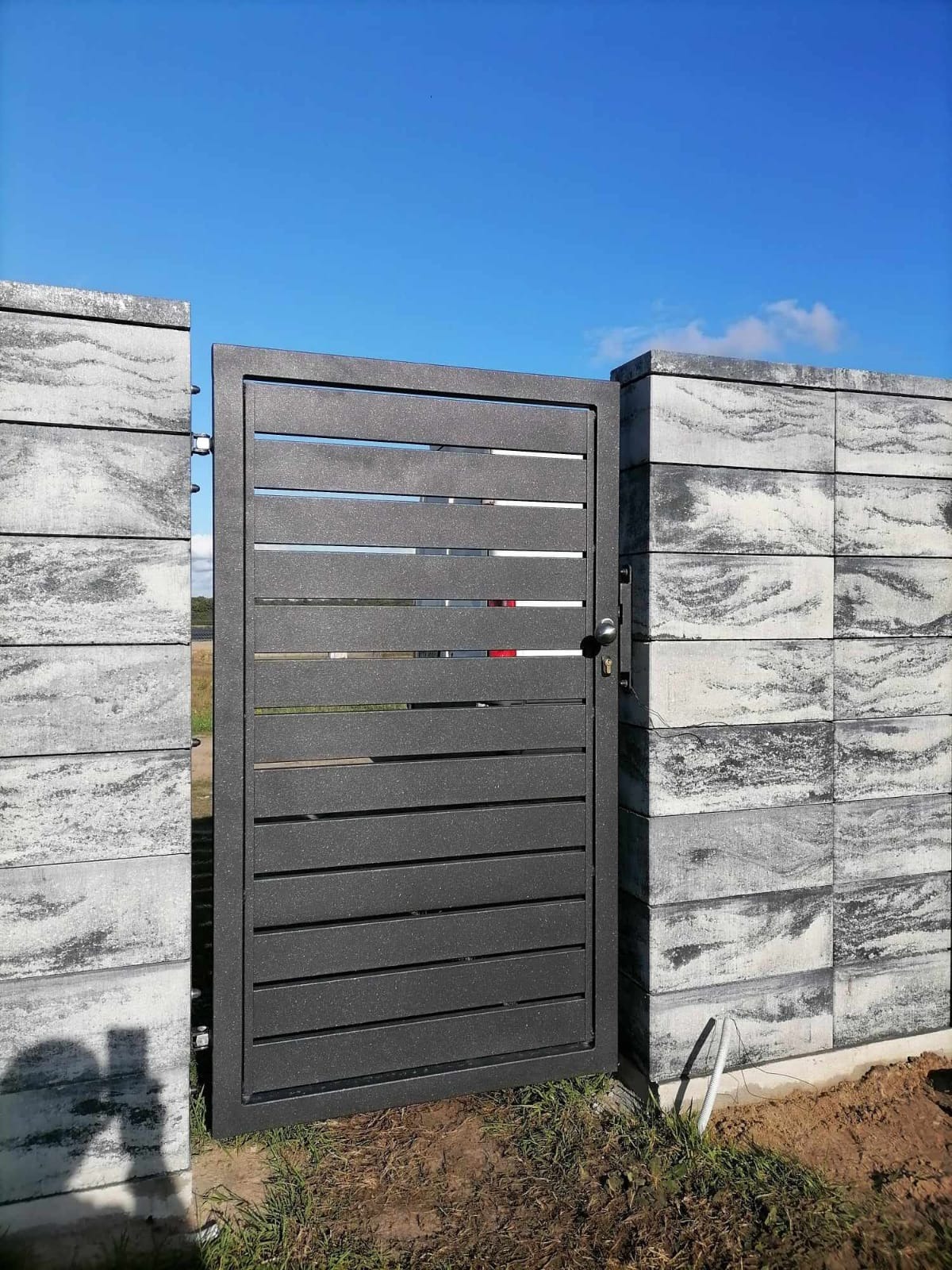 Nowoczesna, grafitowa furtka z poziomych profili, zamontowana w ogrodzeniu z betonowych bloczków imitujących kamień, błękitne niebo w tle.