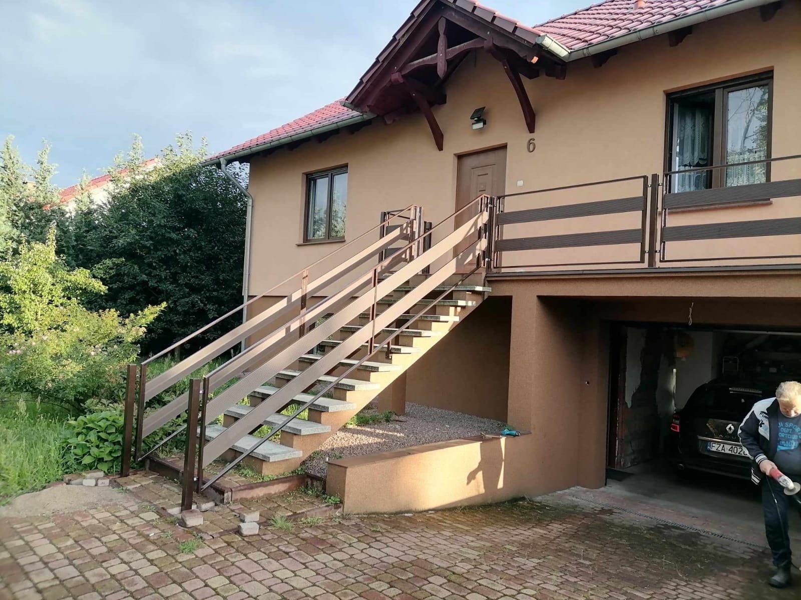 Zewnętrzne schody z brązowymi metalowymi poręczami prowadzące do drzwi wejściowych na piętrze domu z garażem, widoczne elementy balustrady balkonowej.