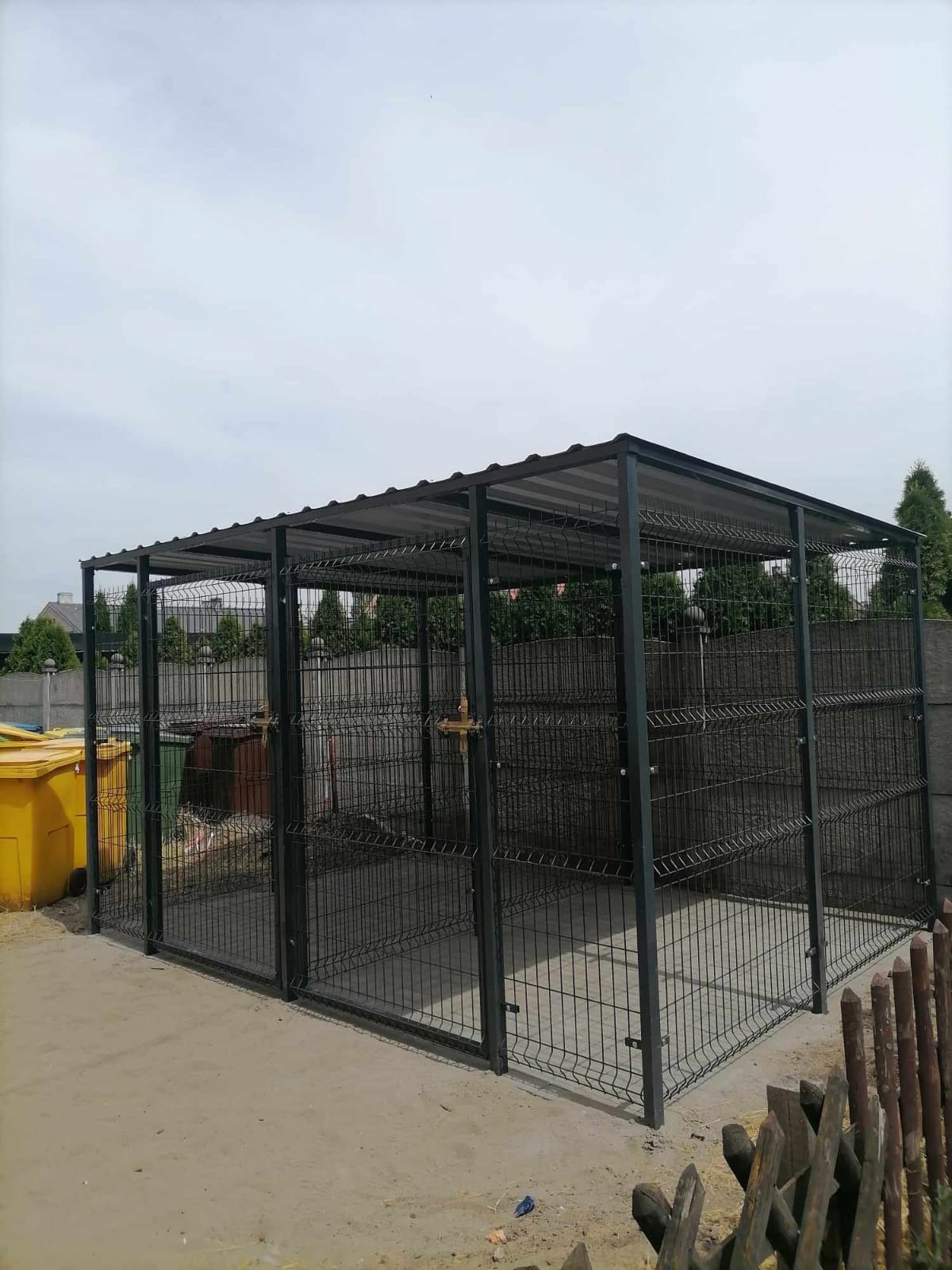 Metalowa konstrukcja z zadaszeniem z paneli ogrodzeniowych, prawdopodobnie kojec dla zwierząt, z żółtymi i zielonymi pojemnikami na śmieci w tle.