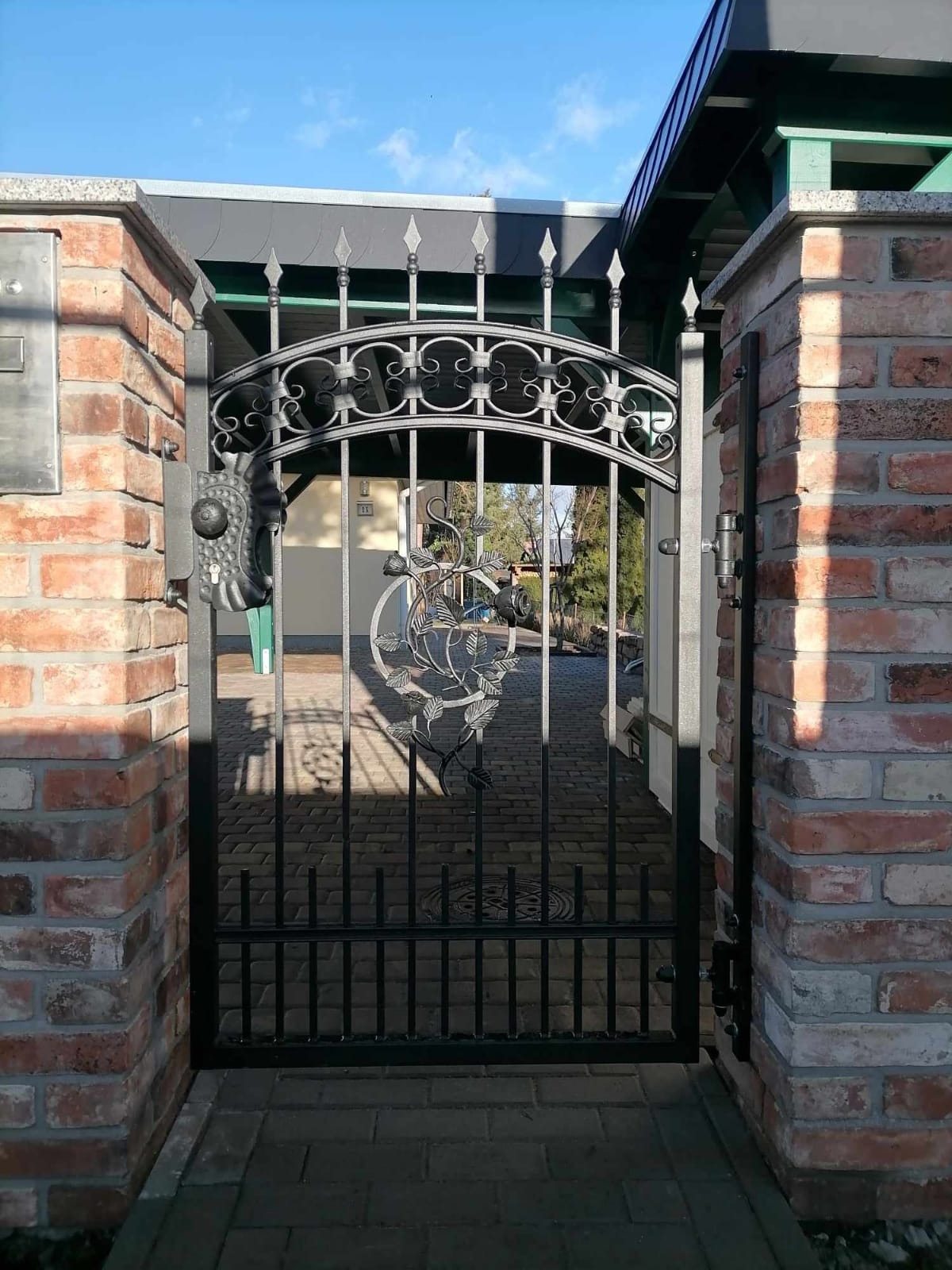 Otwarta, czarna, metalowa furtka z dekoracyjnym motywem roślinnym, osadzona między dwoma murowanymi, ceglanymi słupkami, prowadząca na wybrukowany podjazd.