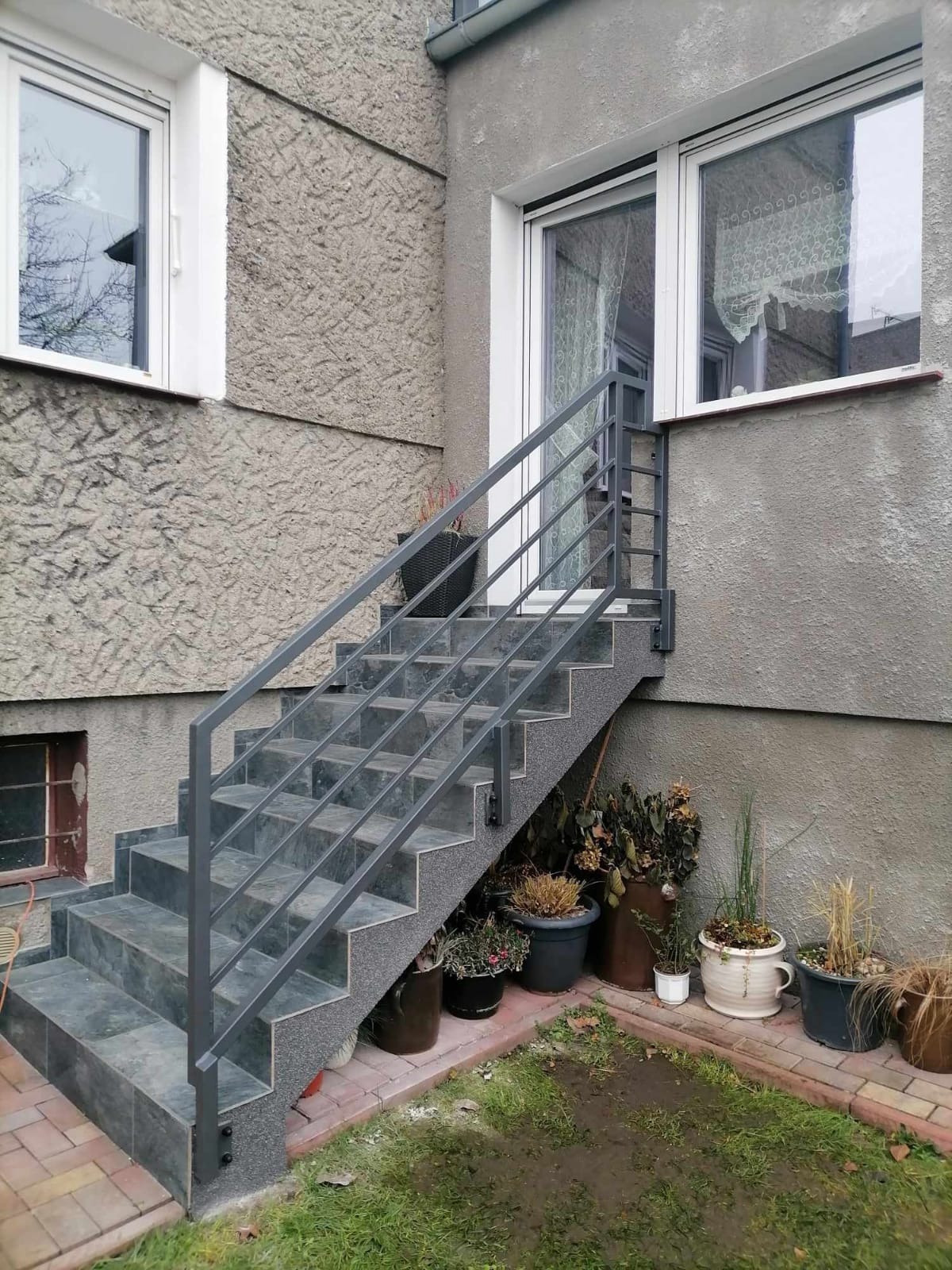 Zewnętrzne schody z metalową poręczą w kolorze szarym, prowadzące do drzwi wejściowych budynku z szarą, chropowatą elewacją, obok schodów ustawione donice z roślinami.