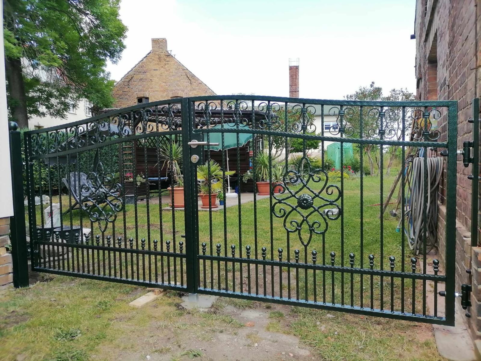 Otwarta, dwuskrzydłowa, metalowa brama z dekoracyjnymi elementami w kształcie rozet, prowadząca do zadbanego ogrodu z domem w tle.