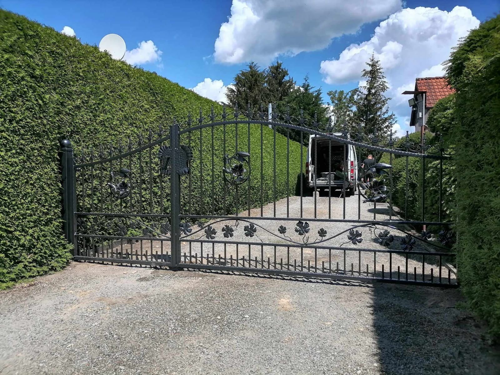 Kuta brama wjazdowa z motywem roślinnym, Lubsko, z widocznym samochodem dostawczym w tle i wysokim żywopłotem po bokach.
