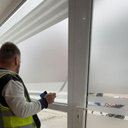 Sebastian Matusik Firma Handlowo-Usługowa - Pracownik w kamizelce odblaskowej aplikuje folię matującą na okno biurowe, używając rakli do wygładzania powierzchni.