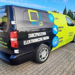 Sebastian Matusik Firma Handlowo-Usługowa - Czarno-żółty samochód dostawczy z reklamą firmy zajmującej się zabezpieczeniami elektronicznymi, prezentujący logo i dane kontaktowe, zaparkowany na brukowanym placu w słoneczny dzień.