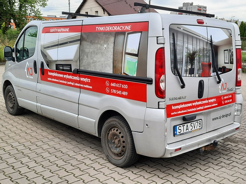 Srebrny samochód dostawczy Renault z reklamą firmy AJ Design oferującej kompleksowe wykończenia wnętrz, w tym sufity napinane i tynki dekoracyjne, zaparkowany na kostce brukowej z widocznymi...