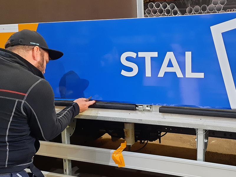 Mężczyzna w czarnej bluzie i czapce, z bliska, przykłada czarną listwę do niebieskiej powierzchni z białym napisem 'STAL' i częścią białego logo, prawdopodobnie podczas montażu na konstrukcji pojazdu.