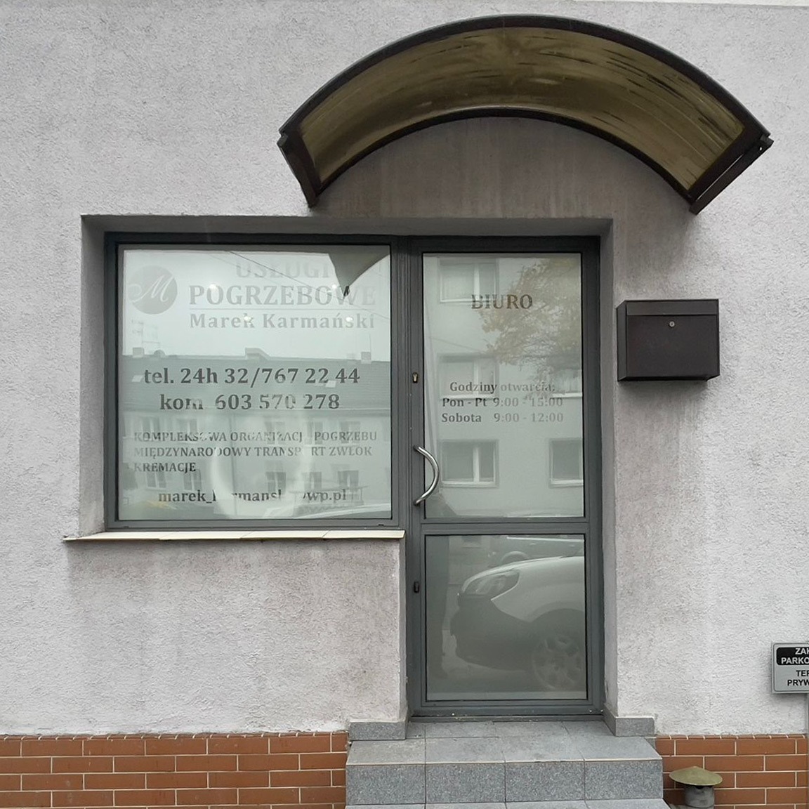 Wejście do biura usług pogrzebowych z naklejonymi informacjami na szybie, widoczna skrzynka pocztowa i zadaszenie nad drzwiami.