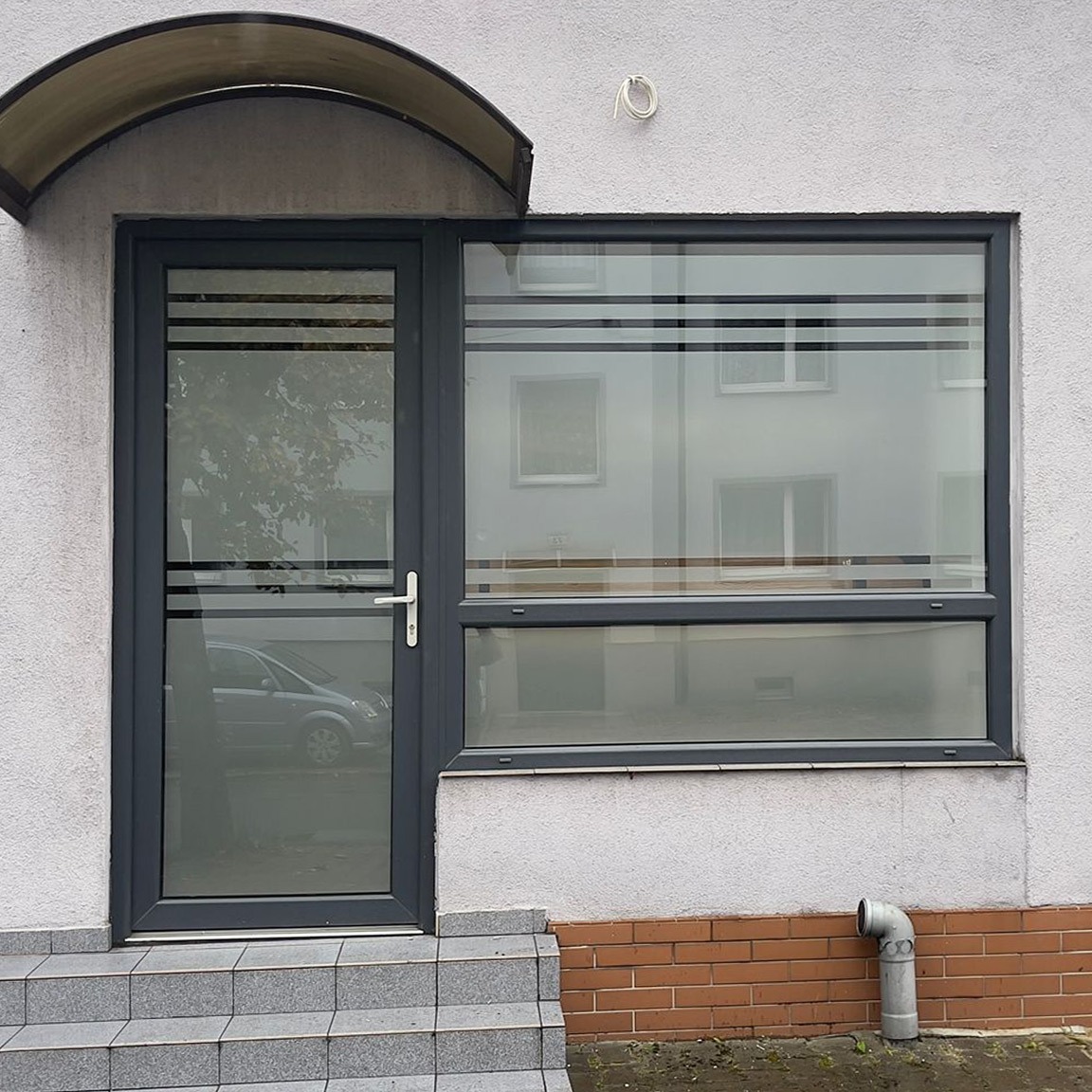 Szare aluminiowe drzwi wejściowe z szybą i przylegające okno z poziomymi paskami, widoczne schody wejściowe i fragment elewacji z rurą odpływową.