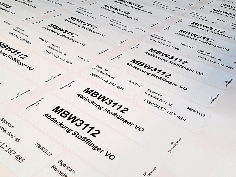 Arkusz z powtarzalnymi, białymi etykietami z czarnym tekstem 'MBW3112 Abdeckung Stoßfänger VO' oraz danymi firmy 'Eigentum Mercedes Benz AG MBW3112 167 484' ułożonymi pod kątem, widoczne z bliska.