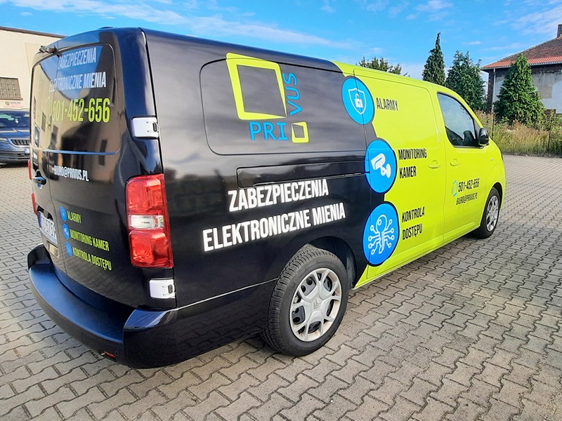 Czarno-żółty samochód dostawczy z reklamą firmy zajmującej się zabezpieczeniami elektronicznymi, prezentujący logo i dane kontaktowe, zaparkowany na brukowanym placu w słoneczny dzień.