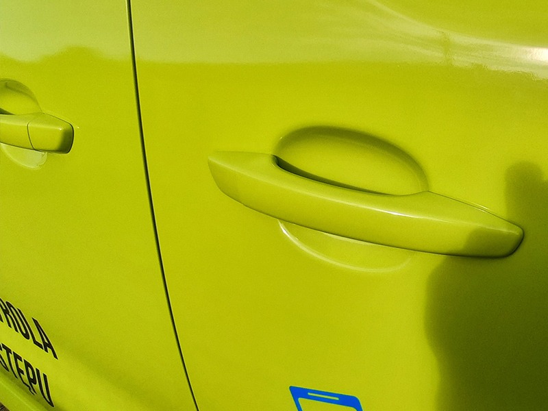 Fragment karoserii samochodu w jaskrawym, limonkowym kolorze z naklejonymi czarnymi napisami i niebieskim logotypem, widoczny uchwyt drzwi.