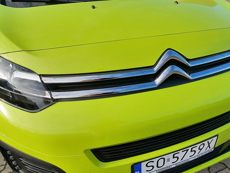 Żółto-zielony Citroen, widok z przodu, zbliżenie na maskę, grill z chromowanymi listwami i logo, oraz fragment reflektora i tablicy rejestracyjnej.