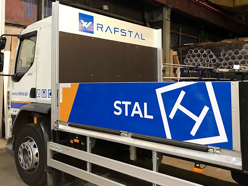 Biała ciężarówka z częściowo oklejoną skrzynią ładunkową, widoczny napis 'STAL' na niebieskim tle z białym logo w kształcie litery H, oraz logo RAFSTAL na górnej części skrzyni.