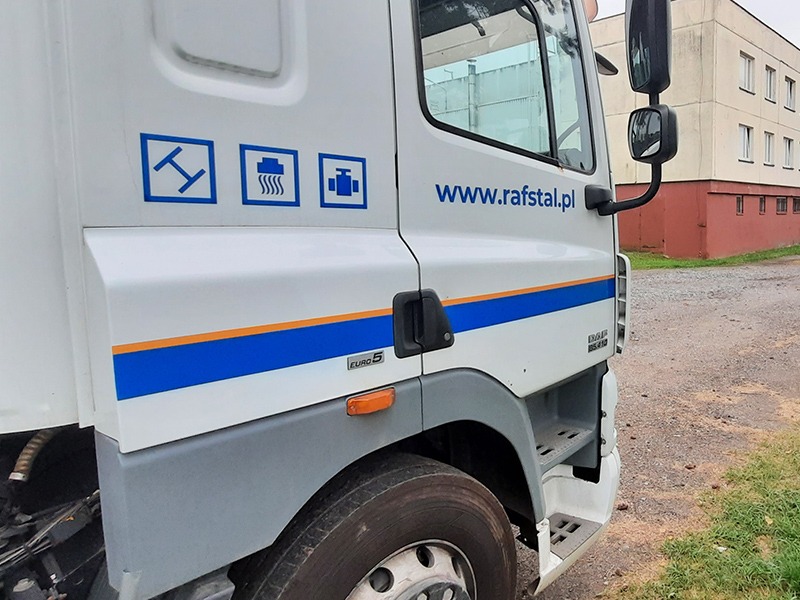 Biała kabina samochodu ciężarowego z niebieskimi elementami graficznymi, w tym logo i adresem strony internetowej www.rafstal.pl, na tle szarego budynku.