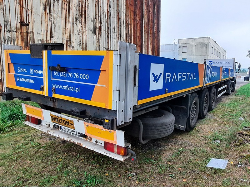 Oklejona ciężarówka z logo Rafstal, numerem telefonu i adresem strony internetowej na tle zardzewiałego budynku.