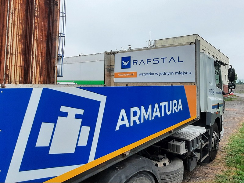 Ciężarówka z niebieskim nadrukiem 'ARMATURA' oraz logo i nazwą firmy Rafstal na białym tle, zaparkowana na żwirowym placu, z widocznym fragmentem zardzewiałego zbiornika w tle.