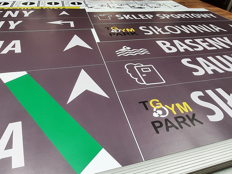 Brązowe tablice informacyjne z białymi napisami i piktogramami, wskazujące kierunek do sklepu sportowego, siłowni, basenów i sauny, z logo TGym Park.
