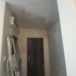 Marcin Hnizdo - Korytarz w trakcie remontu: ściany i sufit po szpachlowaniu, przygotowane do malowania, widoczna drabina aluminiowa i ciemne drzwi.