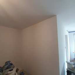 Marcin Hnizdo - Świeżo pomalowany pokój z widocznym narożnikiem ściany i sufitu, z zabezpieczoną taśmą malarską framugą drzwiową i stertą materiałów budowlanych w rogu pomieszczenia.