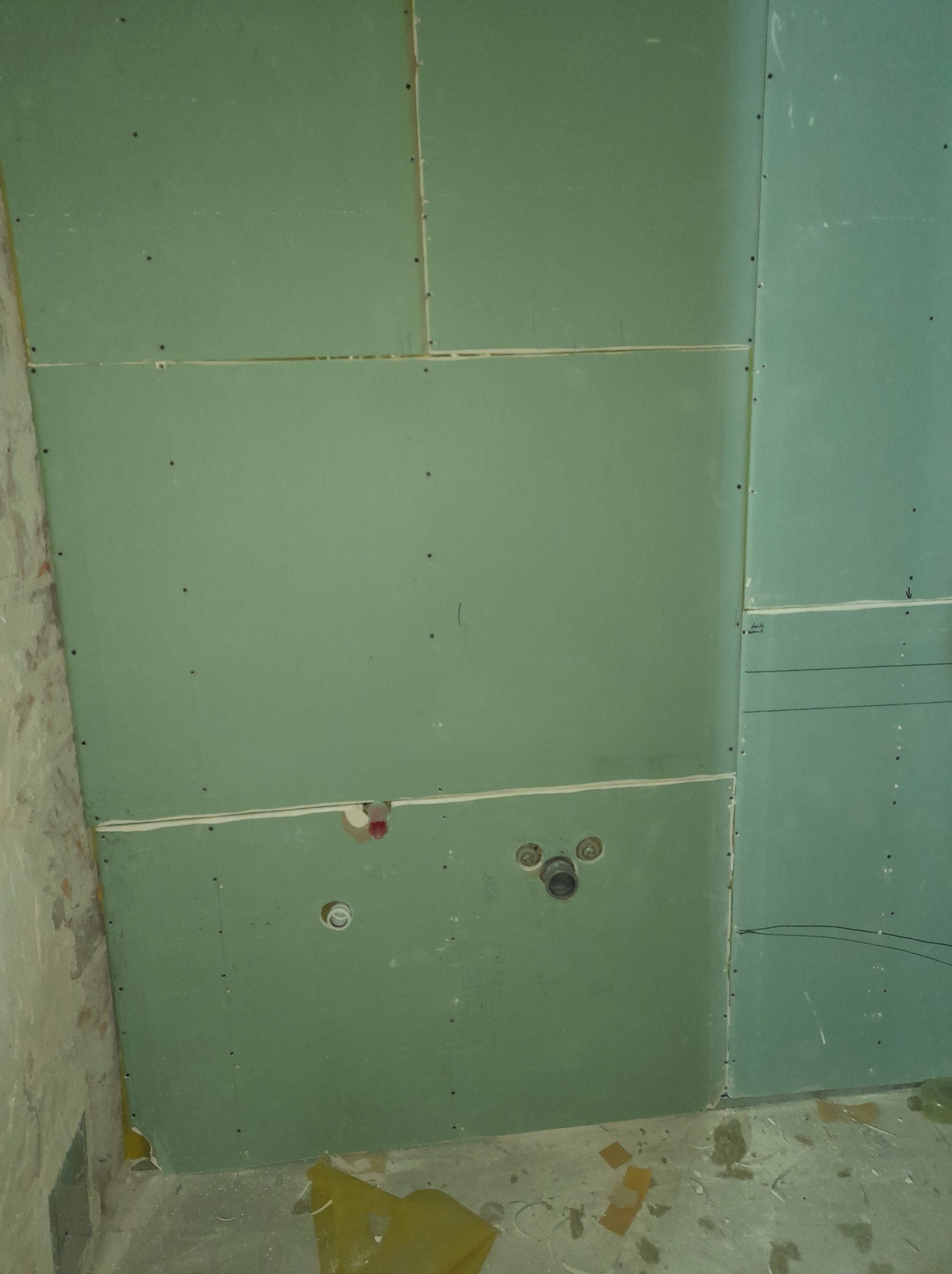 Zielone płyty gipsowo-kartonowe zamontowane na ścianie w łazience, z widocznymi otworami na instalację wodną i ślady po ołówku na jednej z płyt.