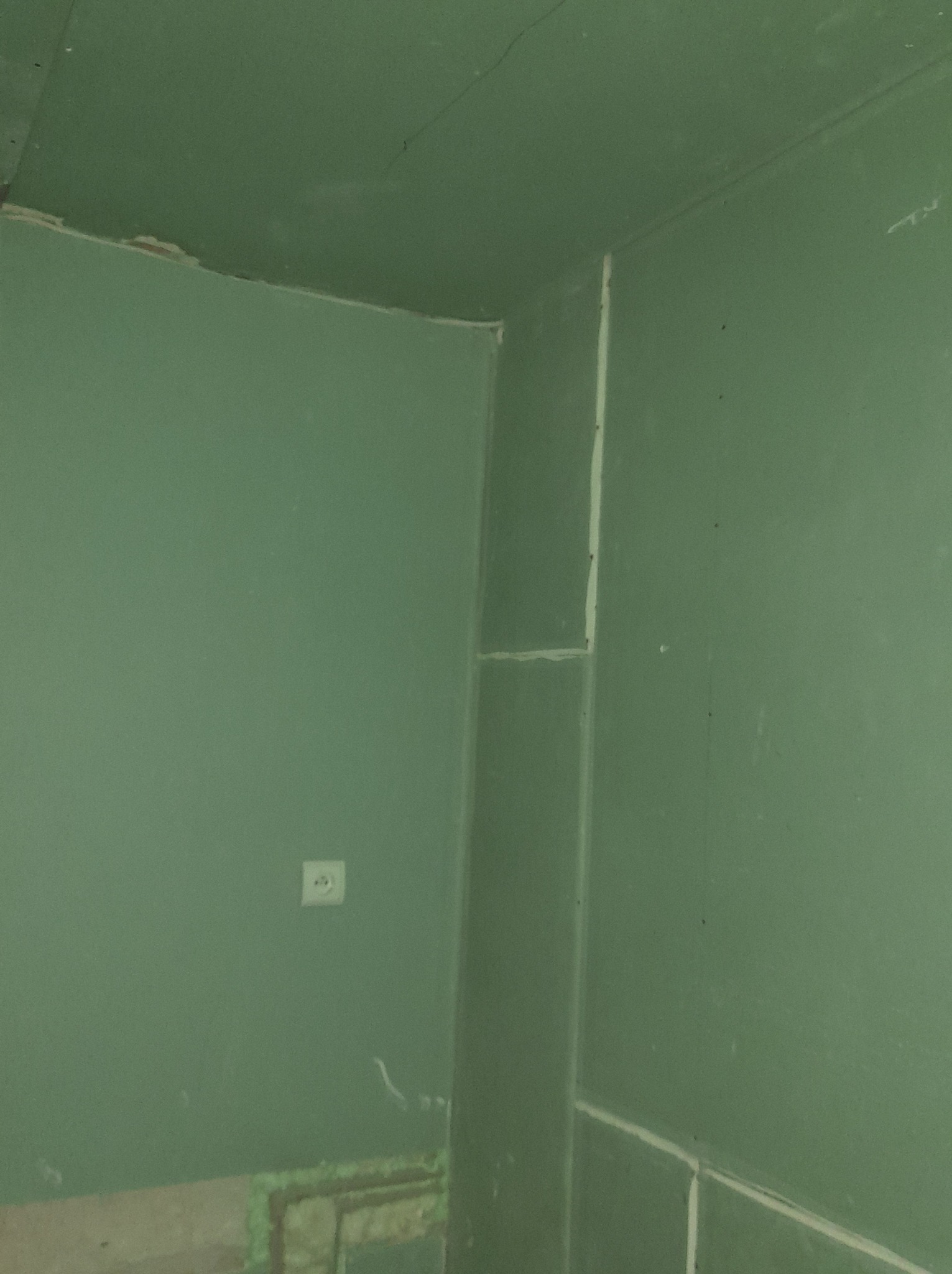 Ściana w rogu pomieszczenia pokryta zielonym podkładem malarskim, widoczne łączenia płyt gipsowo-kartonowych, gniazdko elektryczne i fragment podłogi przed wykończeniem.