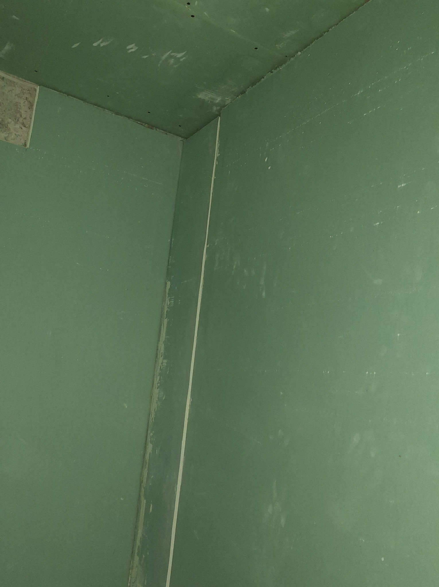 Wewnętrzny róg pomieszczenia z zielonymi płytami kartonowo-gipsowymi, widoczne ślady szpachli na ścianach i suficie, przygotowanie do dalszych prac wykończeniowych.