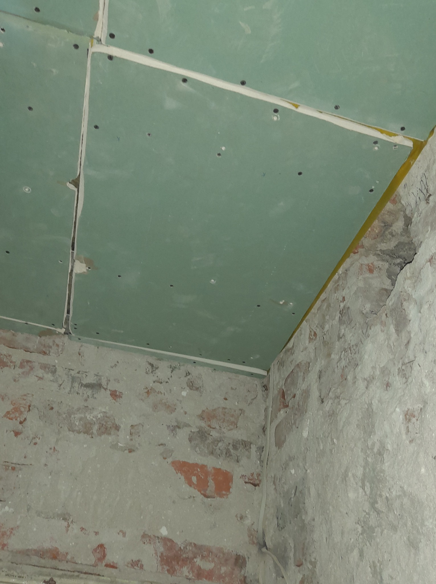 Montaż zielonych płyt na suficie w pomieszczeniu z surową, ceglaną ścianą pokrytą resztkami tynku. Widoczne łączenia płyt z żółtą taśmą, mocowane wkrętami.