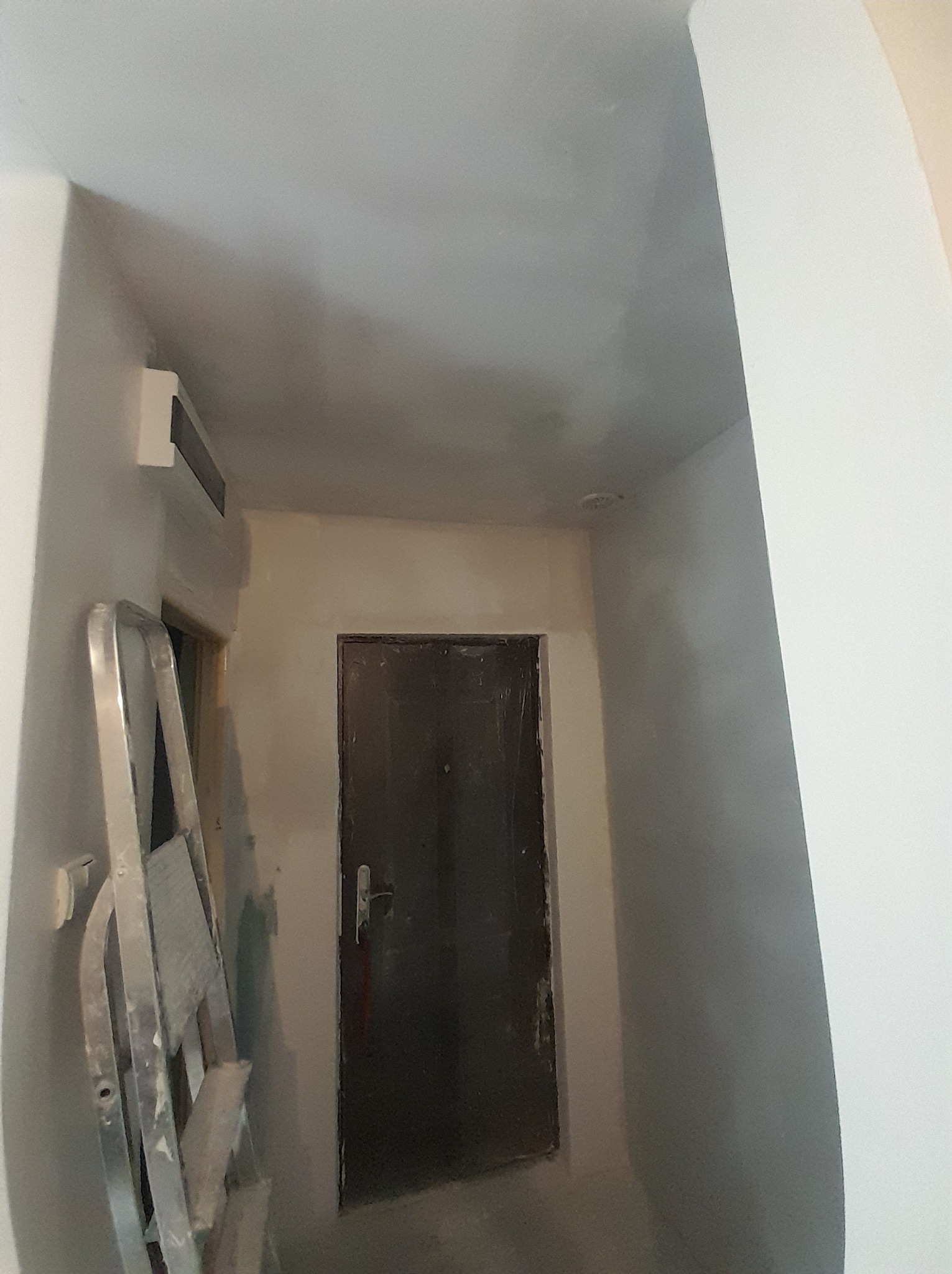 Korytarz w trakcie remontu: ściany i sufit po szpachlowaniu, przygotowane do malowania, widoczna drabina aluminiowa i ciemne drzwi.