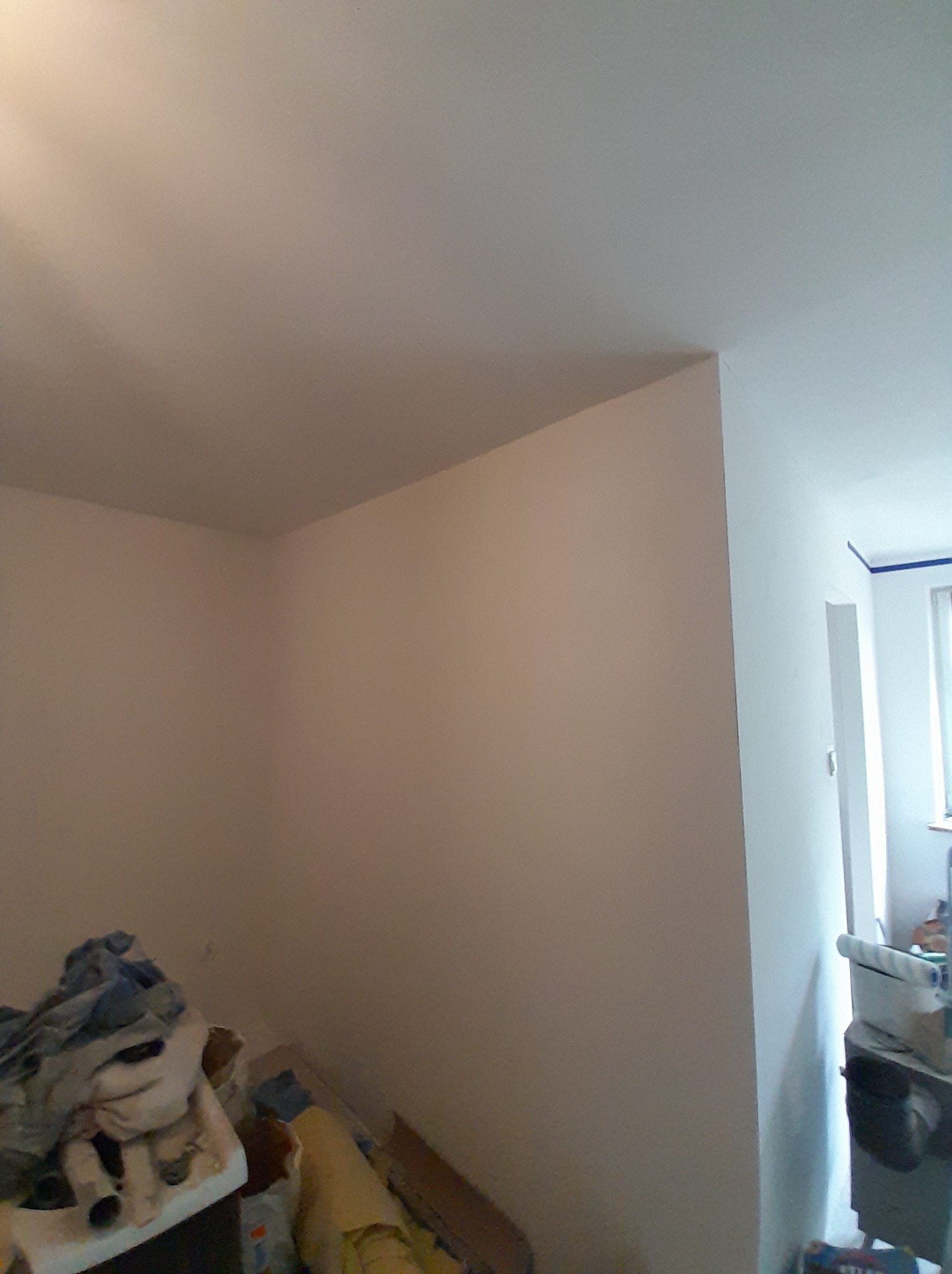 Świeżo pomalowany pokój z widocznym narożnikiem ściany i sufitu, z zabezpieczoną taśmą malarską framugą drzwiową i stertą materiałów budowlanych w rogu pomieszczenia.