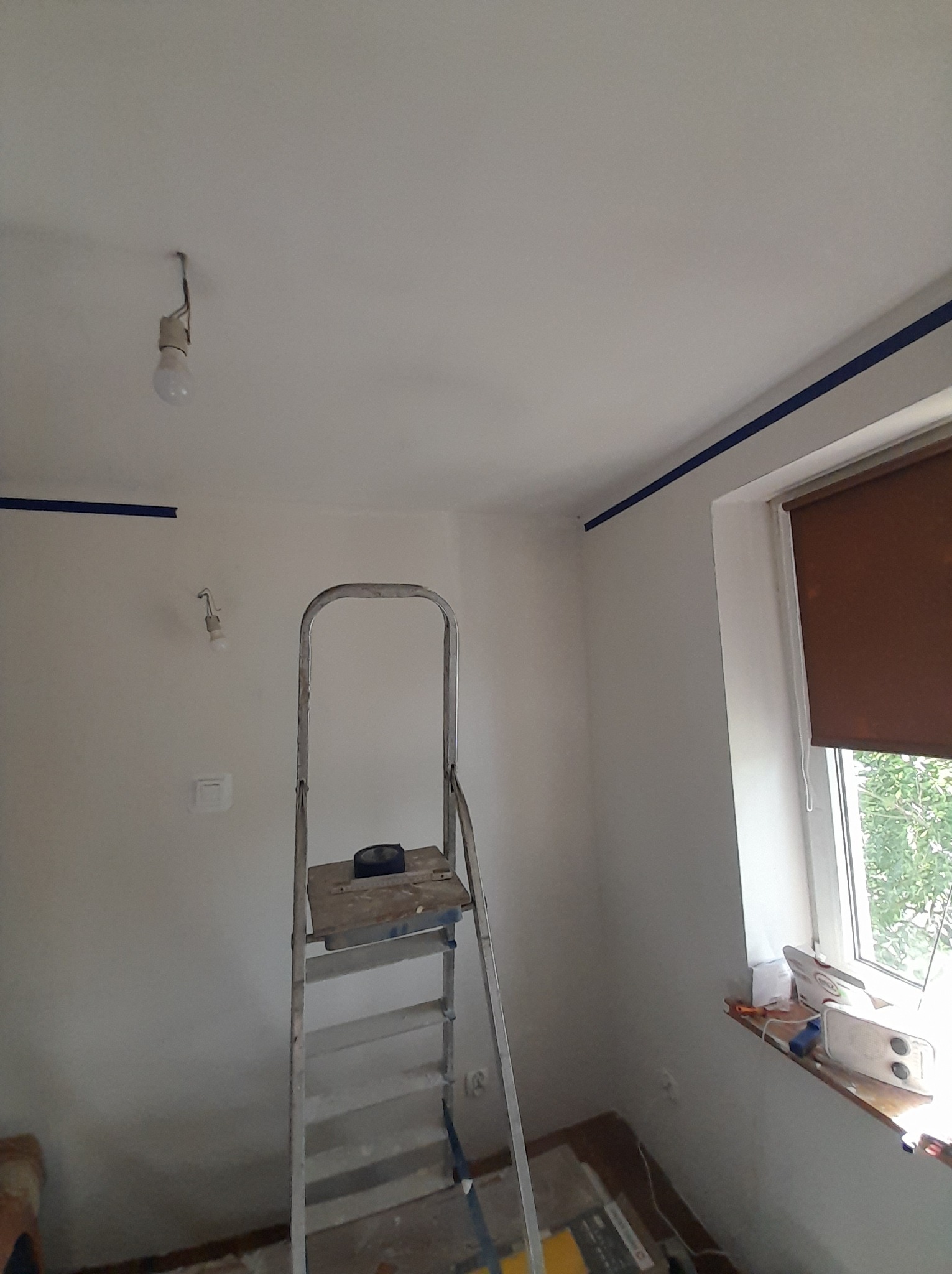 Pokój w trakcie malowania: ściany pomalowane na biało, zabezpieczone niebieską taśmą malarską przy suficie i oknie, drabina aluminiowa w centrum, żarówka na kablu zwisająca z sufitu.