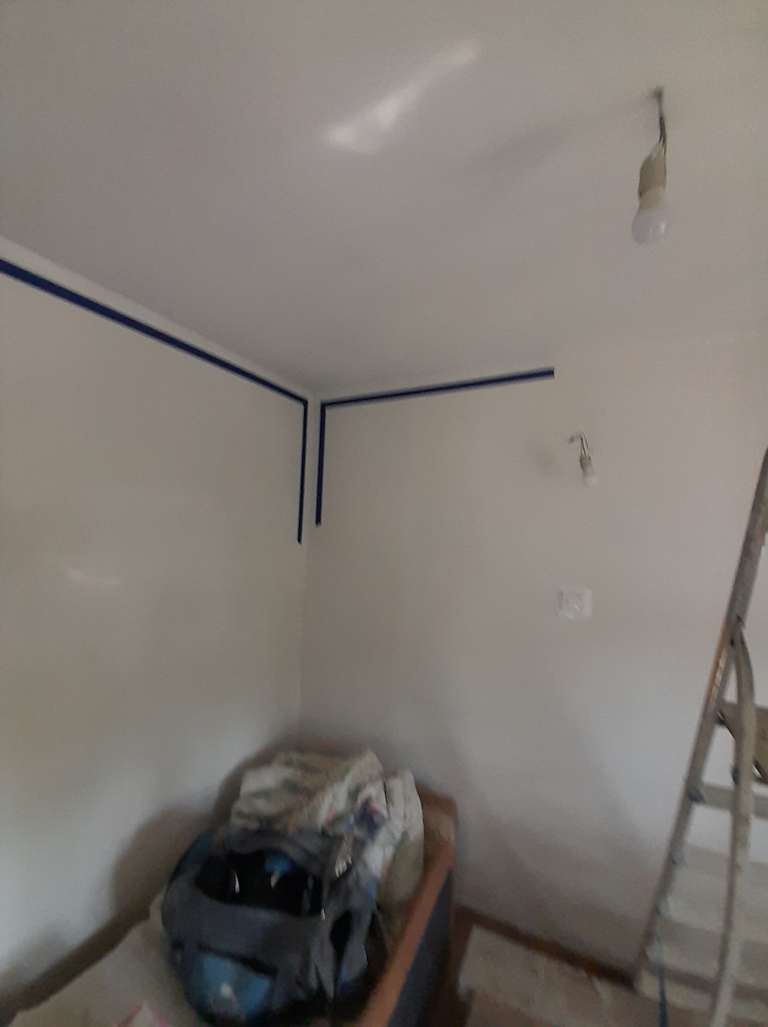 Przygotowanie do malowania ścian: widoczne taśmy malarskie przy suficie, drabina oparta o ścianę, zakryte meble i wisząca żarówka.