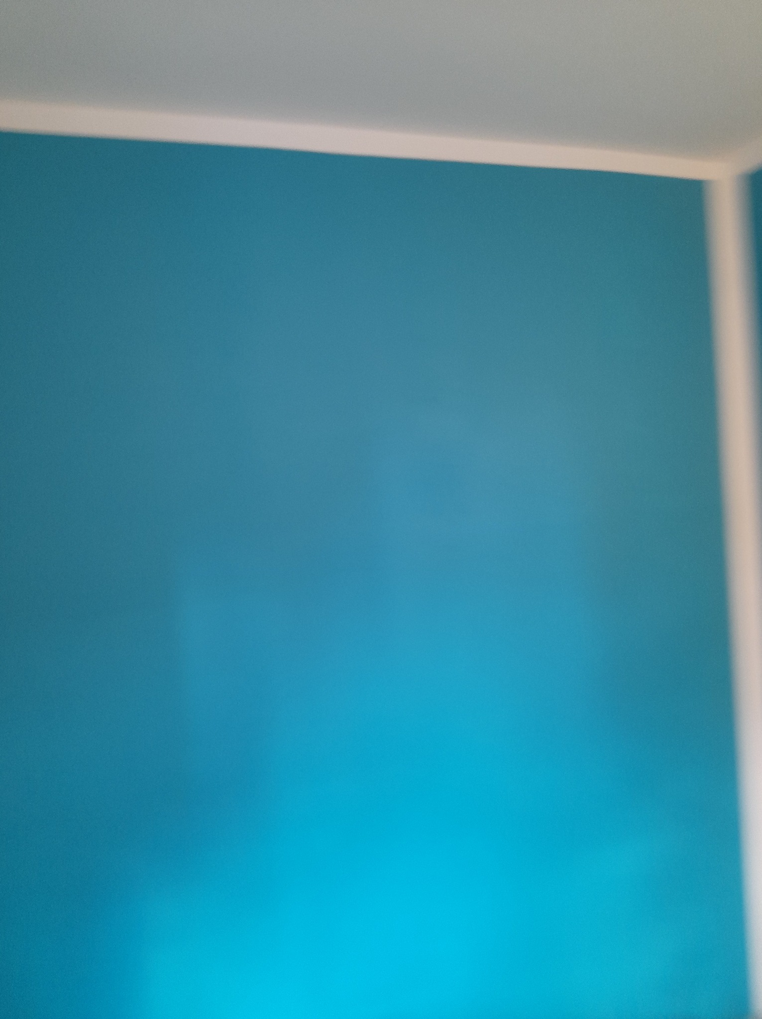 Ściana pomalowana na turkusowo z białą listwą przypodłogową i sufitową, widoczny fragment narożnika pokoju, nierównomierne oświetlenie