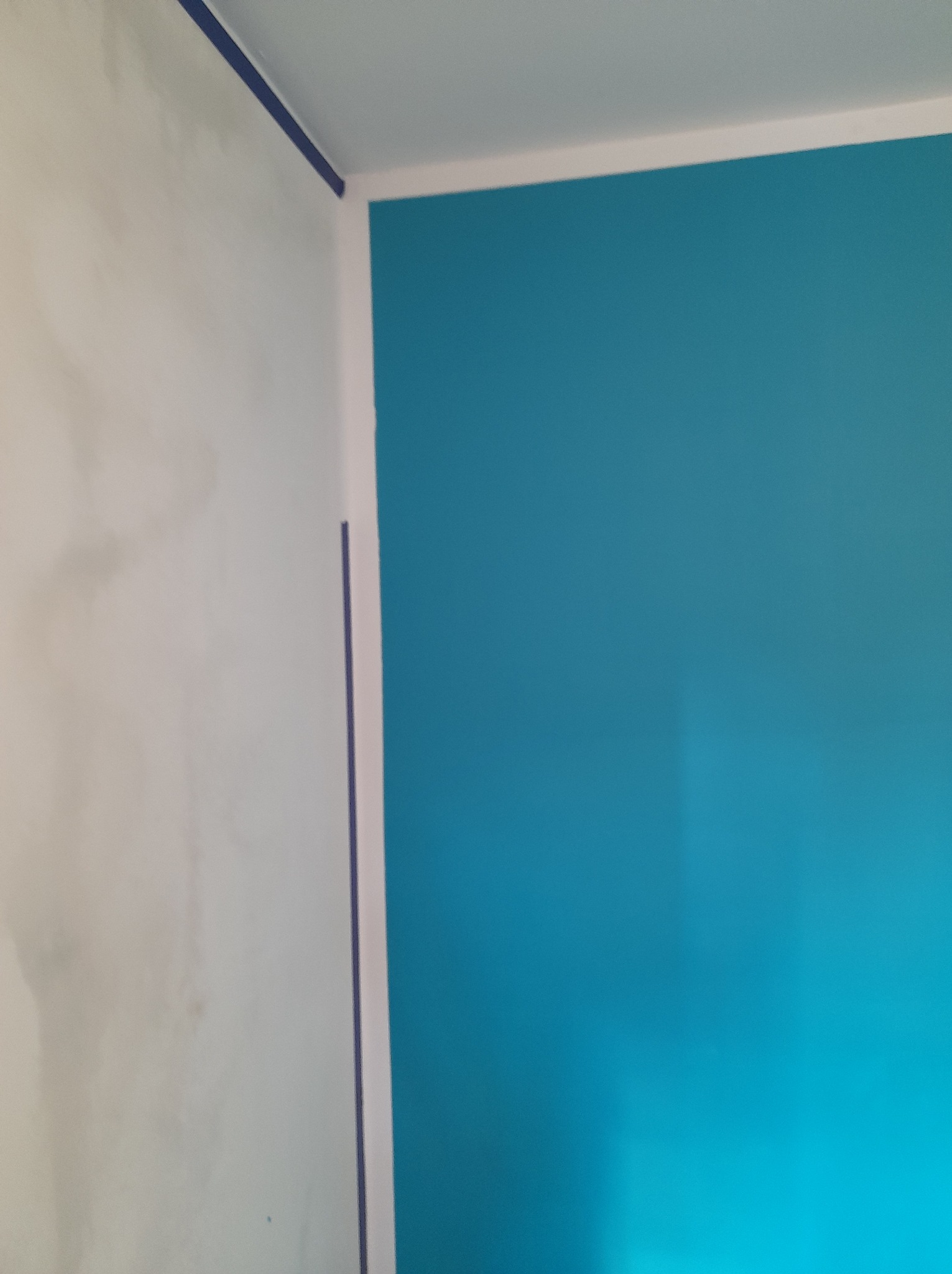 Ściana po szpachlowaniu, z nałożoną taśmą malarską i pomalowana na turkusowo, widoczne niedoskonałości szpachli na sąsiedniej ścianie.