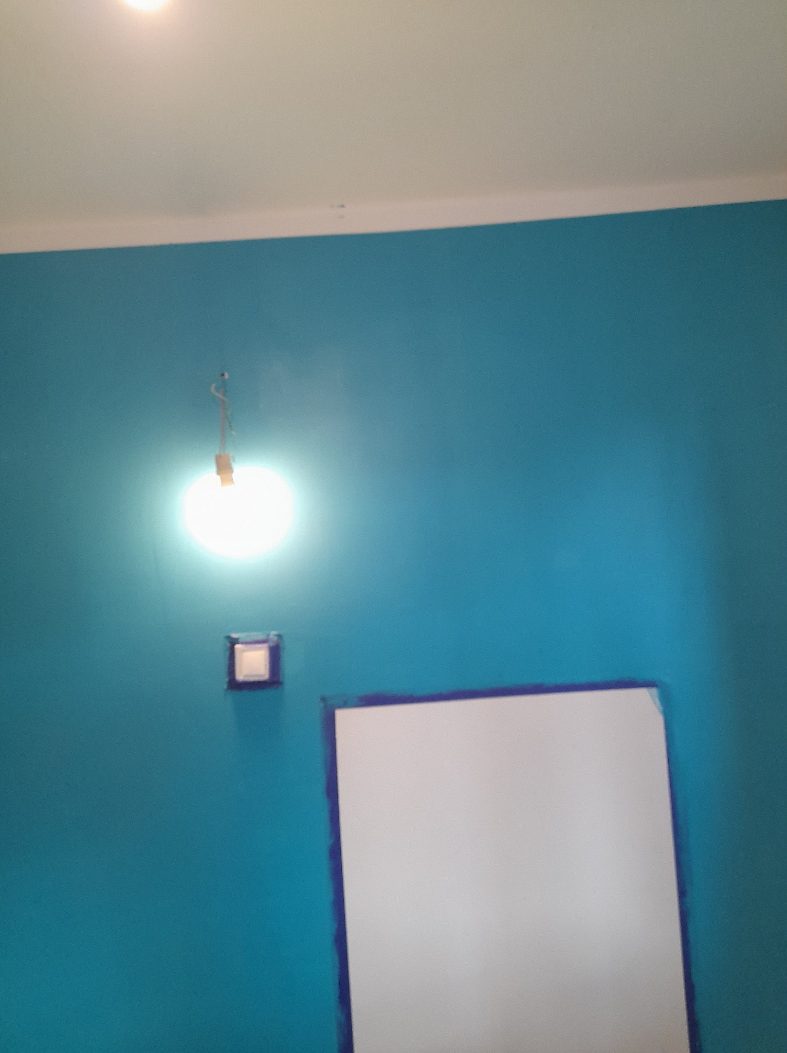 Świeżo pomalowana turkusowa ściana z zabezpieczonym białym prostokątnym obszarem, widoczne niedociągnięcia malarskie przy krawędziach, gniazdko elektryczne i wisząca żarówka.