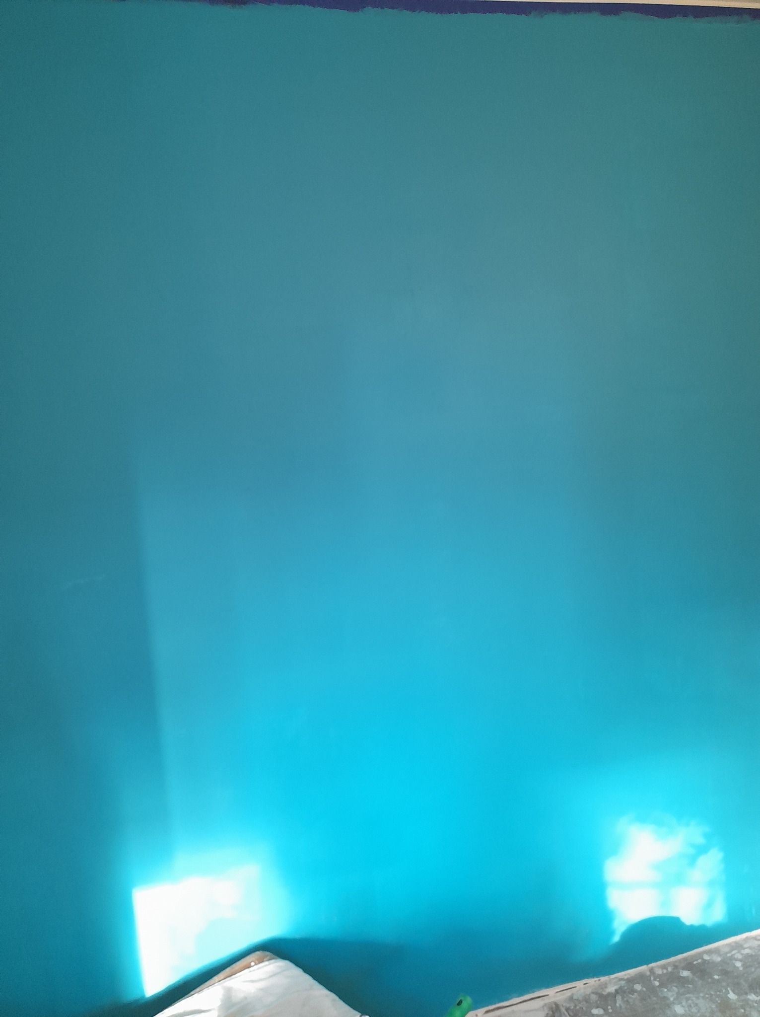 Turkusowa ściana po malowaniu z widoczną taśmą malarską przy suficie i odbiciem światła z okna w dolnej części, widoczna krawędź zabezpieczonej podłogi.