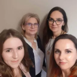 Cztery kobiety, prawdopodobnie zespół biura, pozują do zdjęcia w pomieszczeniu o neutralnym tle.