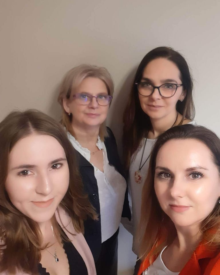 Cztery kobiety, prawdopodobnie zespół biura, pozują do zdjęcia w pomieszczeniu o neutralnym tle.