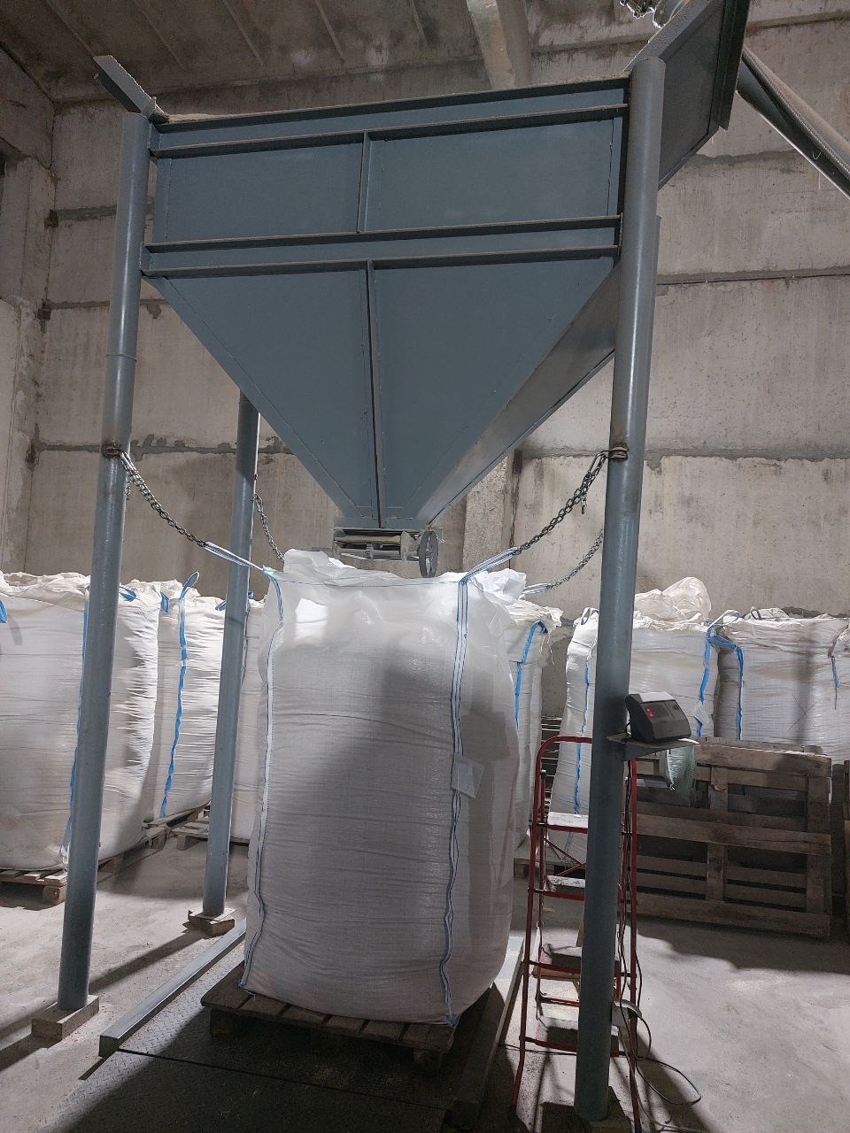 Magazyn z workami typu big bag pod metalową konstrukcją z lejkiem, w tle betonowe ściany i drewniane palety.