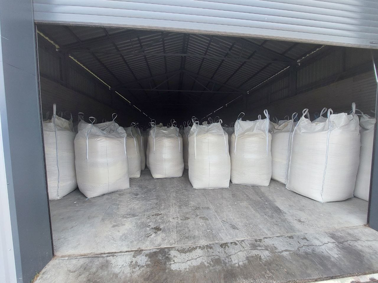 Magazyn z otwartą bramą rolowaną, wypełniony rzędami dużych, białych worków z zawiązanymi sznurkami u góry, ustawionych na betonowej podłodze.