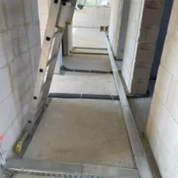 Wnętrze w surowym stanie z widocznymi korytami kablowymi na podłodze, drabina oparta o ścianę, przewody w peszlach. Widoczne puszki elektryczne w ścianach.