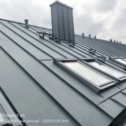 Szary metalowy dach z oknami dachowymi, kominem i odpowietrznikami, widok z dołu pod kątem, pochmurne niebo w tle.