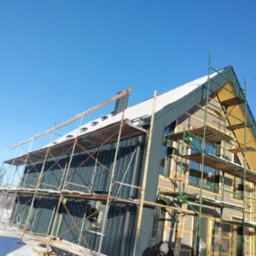 Budynek w trakcie prac wykończeniowych elewacji, widoczne rusztowania i częściowo zamontowana blacha na ścianach, ocieplenie poddasza w trakcie montażu, zimowy krajobraz w tle.