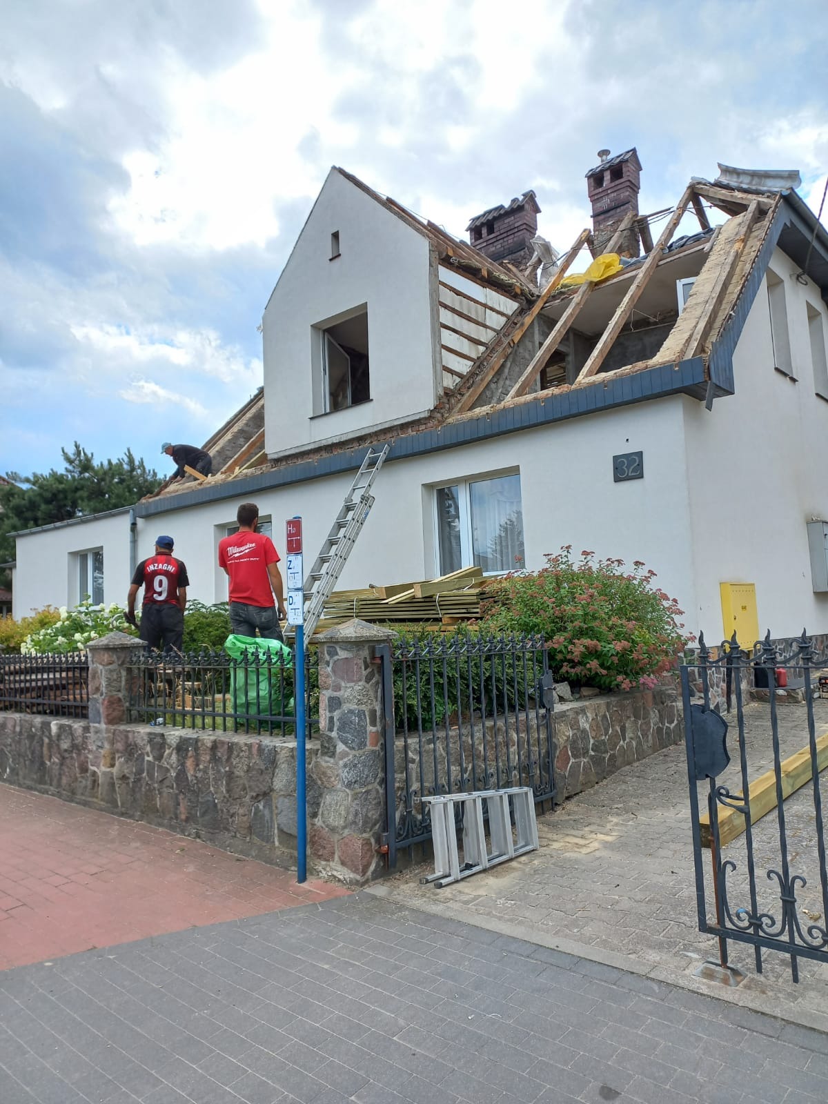 Dom jednorodzinny z częściowo zdemontowanym dachem, widoczne krokwie i ekipa remontowa na dachu oraz na posesji, przygotowane deski na wymianę pokrycia.