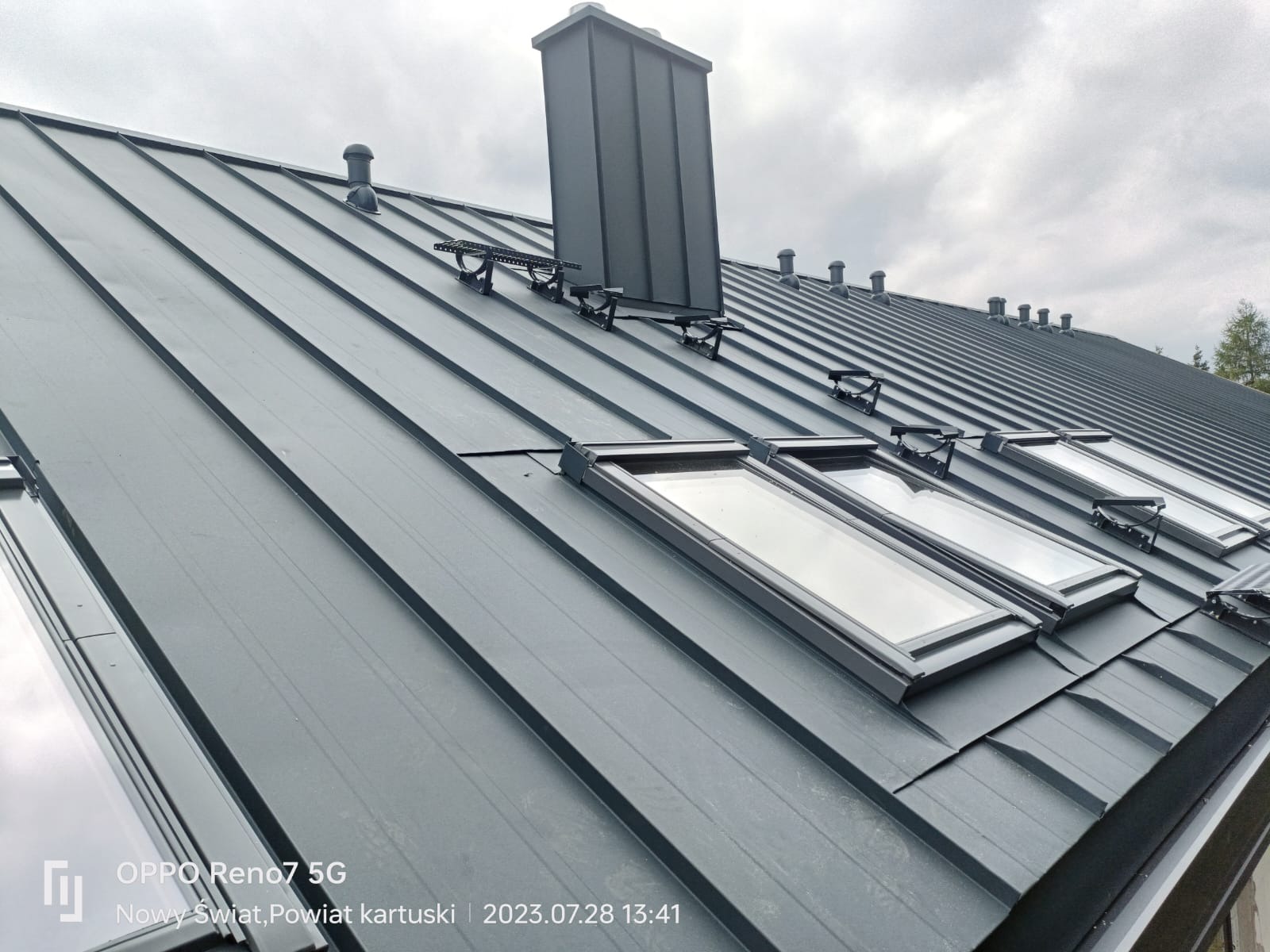 Szary metalowy dach z oknami dachowymi, kominem i odpowietrznikami, widok z dołu pod kątem, pochmurne niebo w tle.