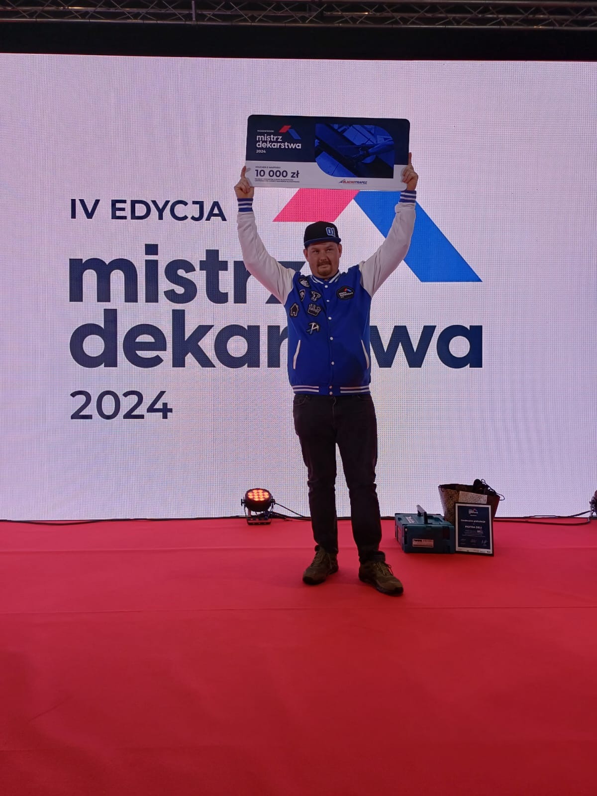 Mężczyzna w bluzie bejsbolówce i czapce z daszkiem, stojący na czerwonej scenie z podniesionymi rękami, trzymając czek na 10 000 zł z napisem 'Mistrz Dekarstwa 2024', obok skrzynka narzędziowa...